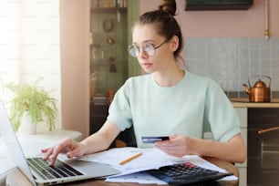 Closeup photo of young European Caucasian girl staying on her kitchen in afternoon holding credit hard and using laptop in order to process online payment enjoying benefits of electronic commerce