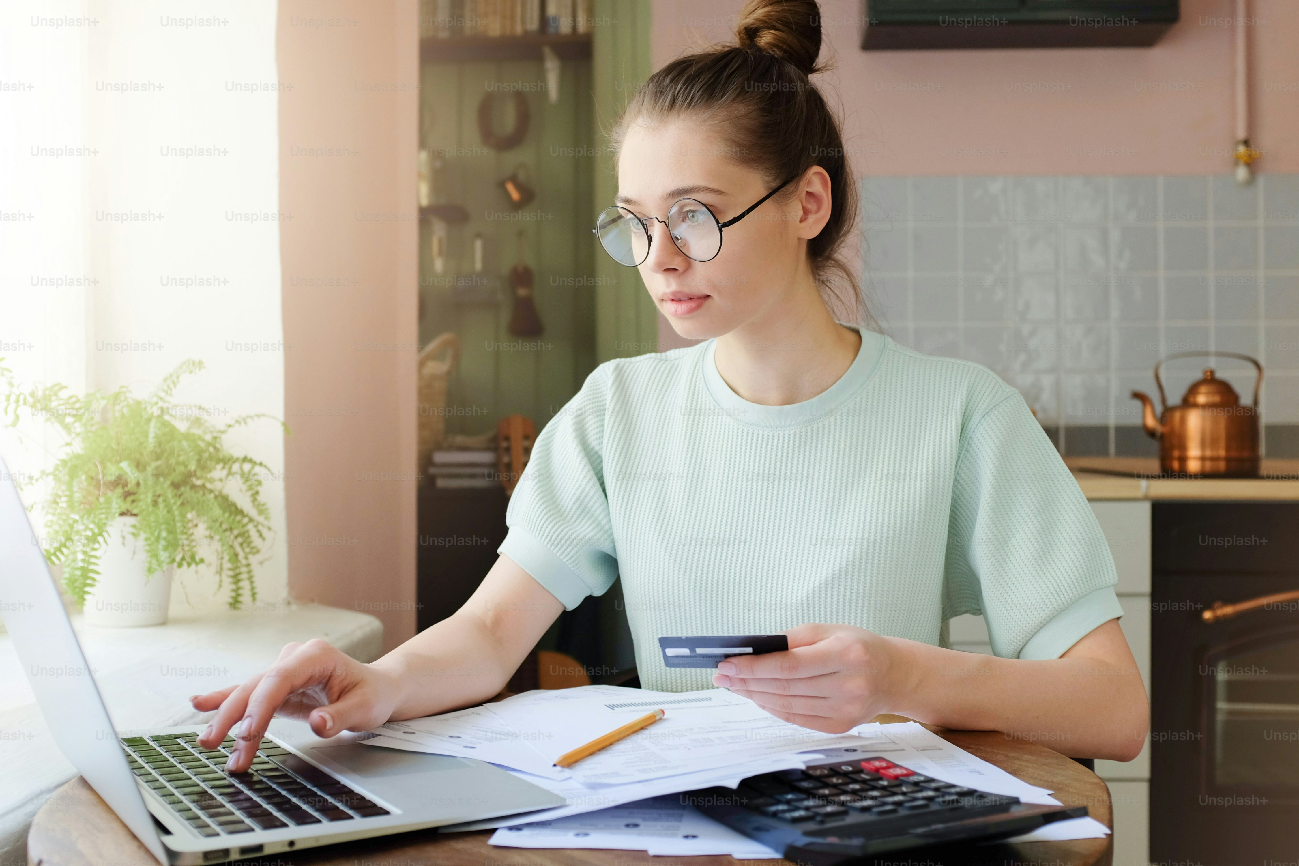 Closeup photo of young European Caucasian girl staying on her kitchen in afternoon holding credit hard and using laptop in order to process online payment enjoying benefits of electronic commerce