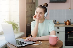 Closeup photo of young good-looking European woman dressed in casual top sitting at kitchen table with white earphones on enjoying favorite audio tracks from laptop with happy smile in free time