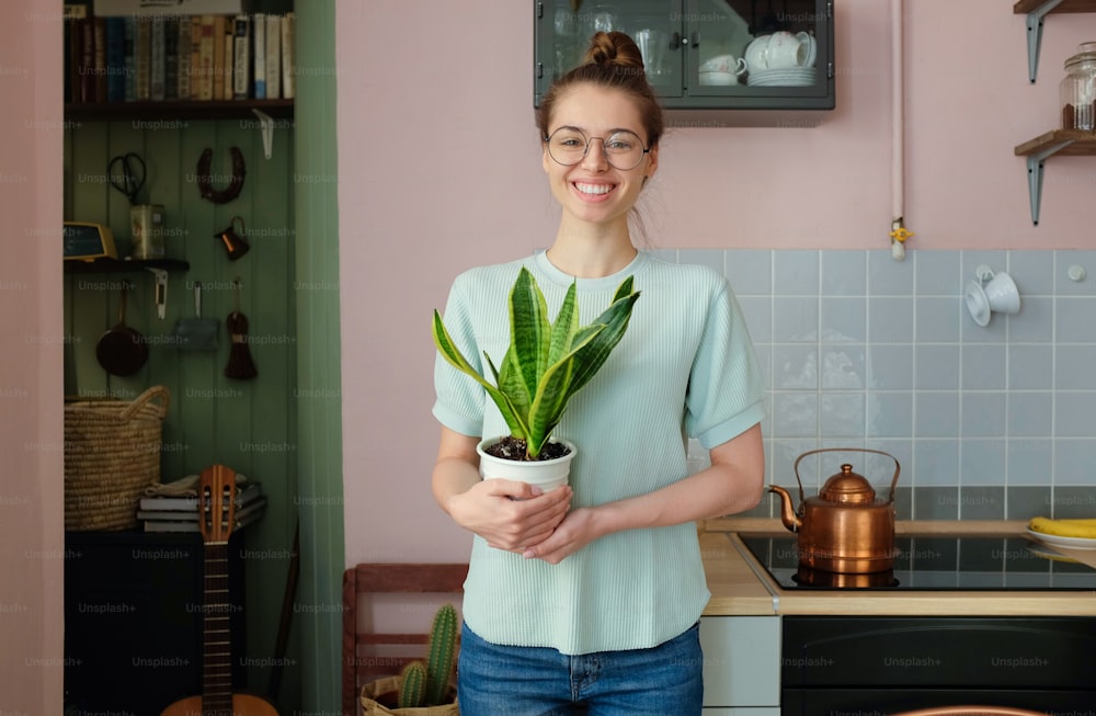 Closeup portrait of attractive Caucasian girl pictured at home wearing T-shirt, jeans and trendy eyeglasses holding potted plant with big green leaves as if having recently moved house, feeling happy
