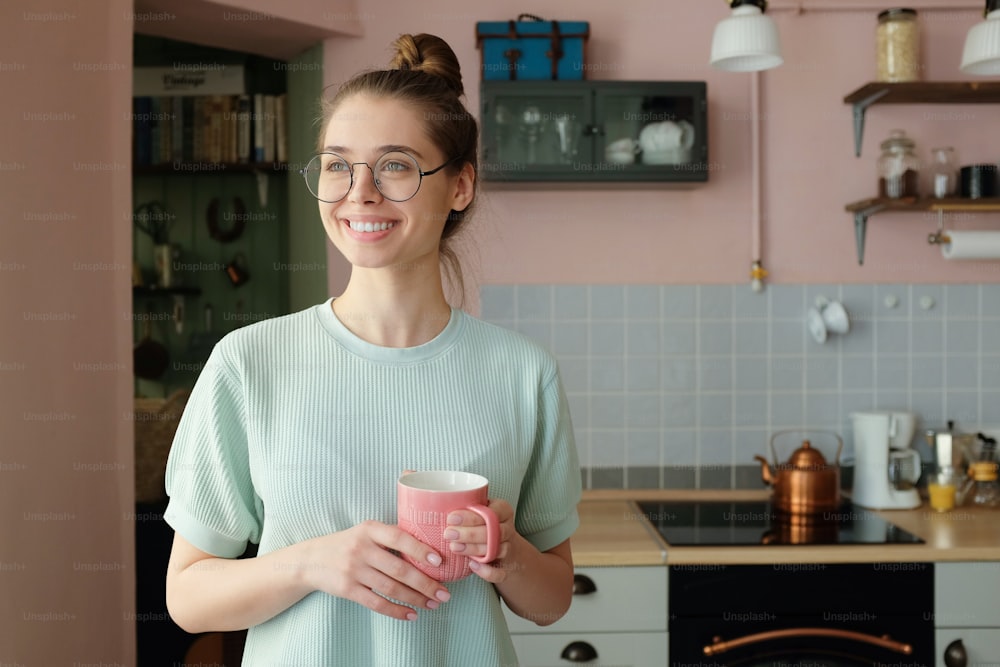 Primer plano de una joven caucásica europea optimista que pasa la tarde en su cocina sosteniendo una taza de bebida en las manos con mirada soñadora, anticipando un buen día fructífero en el trabajo o en casa
