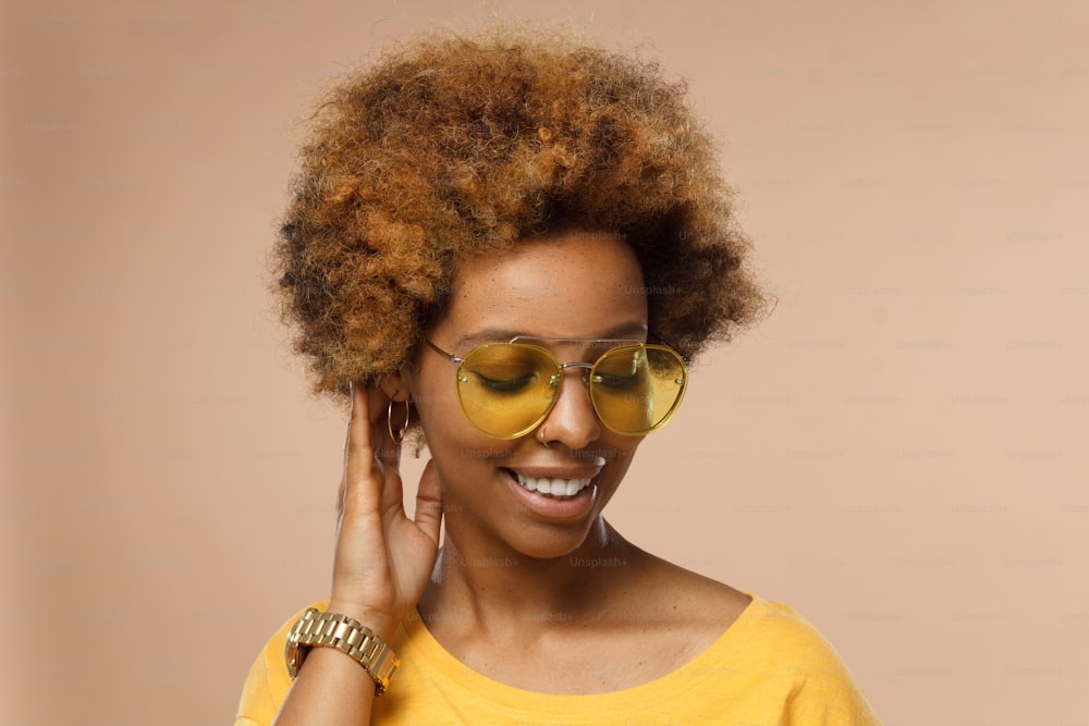 Primo piano orizzontale della giovane donna afroamericana sorridente che flirta pelle scura in occhiali dorati, isolata su sfondo marrone, con la testa girata e toccando i capelli