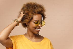 Horizontal photo of young African American girl isolated on beige background dressed in bright yellow top and wearing yellow sunglasses touching her head as if moving to sounds of favorite music