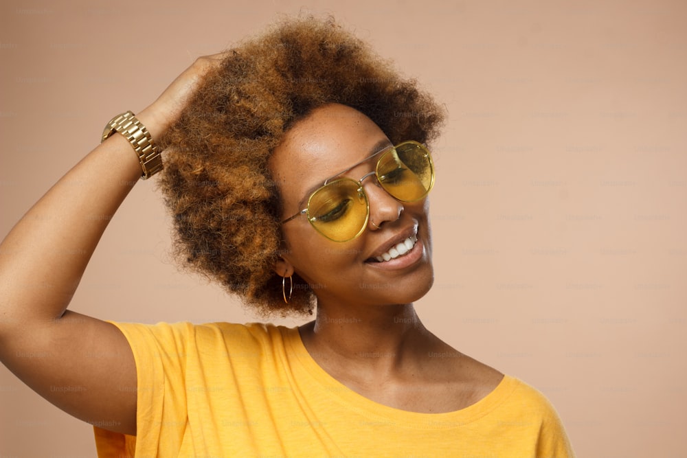 Studio closeup of attractive dark-skinned woman pictured against beige background wearing sunglasses and wristwatch looking down with charming smile as if dreaming or thinking about nice moments