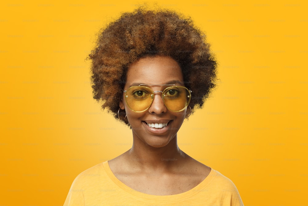 Closeup headshot of African American femalein bright yellow top and wearing glasses of matching color, smiling, ready for having fun with friends
