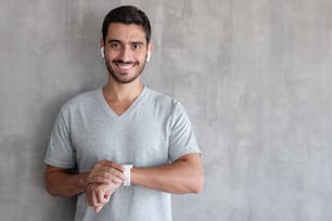Junger lächelnder gutaussehender Mann mit drahtlosen Kopfhörern und T-Shirt, der Smartwatches mit Touchscreen hält und an einer grauen strukturierten Wand steht