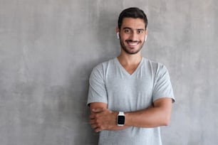 Porträt eines lächelnden, gutaussehenden Mannes in T-Shirt und Smartwatches, der mit verschränkten Armen vor einer grau strukturierten Wand mit Kopierraum steht