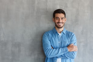Retrato do homem bonito sorridente na camisa azul em pé com os braços cruzados contra a parede texturizada cinza com espaço de cópia