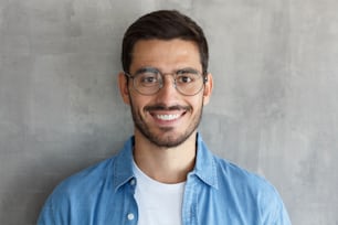 Close up portrait of smiling handsome man in round glasses and blue shirt isolated on gray textured wall