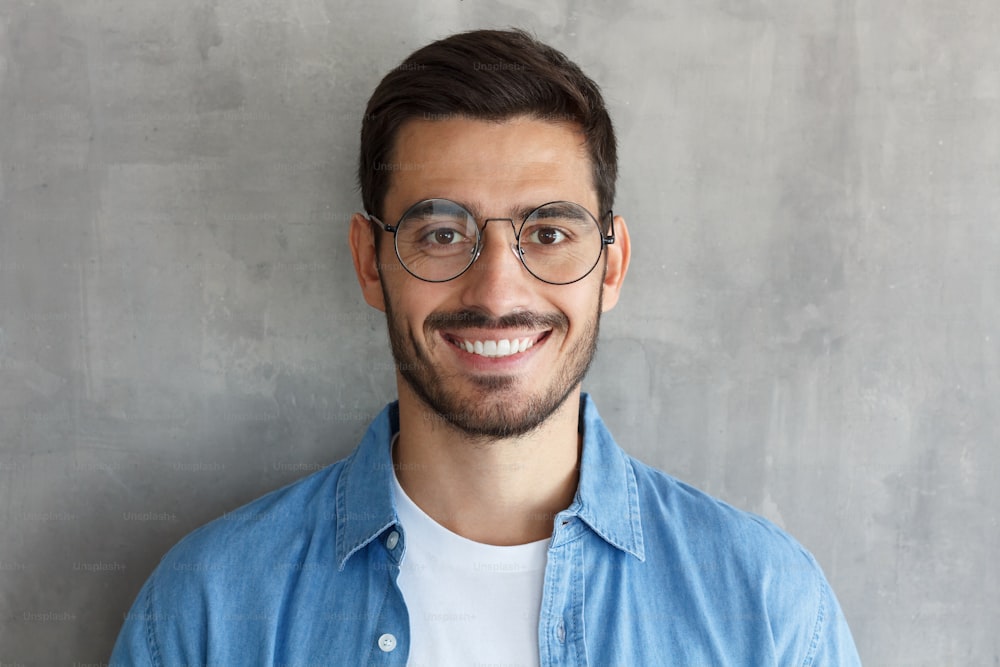 Close up portrait of smiling handsome man in round glasses and blue shirt isolated on gray textured wall