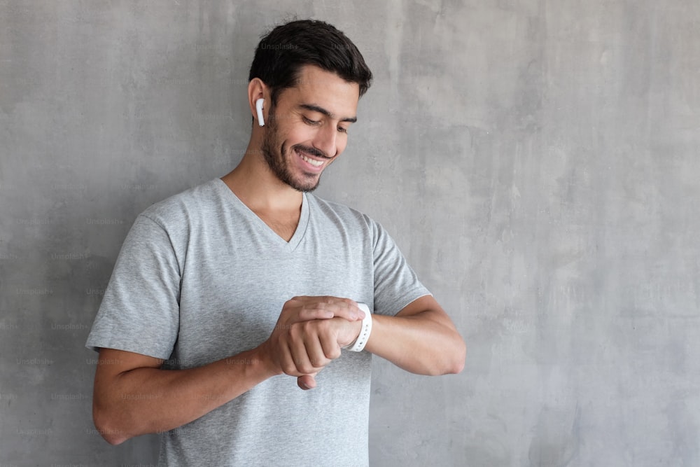 Indoor picture of young European male pictured isolated on grey background enjoying favorite audio tracks through earphones and touching screen of smart watch checking time or adjusting settings