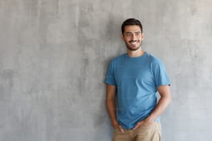 Innenbild eines gutaussehenden kaukasischen Mannes, der isoliert auf grauem Hintergrund vor grauer strukturierter Wand posiert und glücklich lächelt, Copyspace auf der linken Seite, um Produkte für den elektronischen Handel zu bewerben
