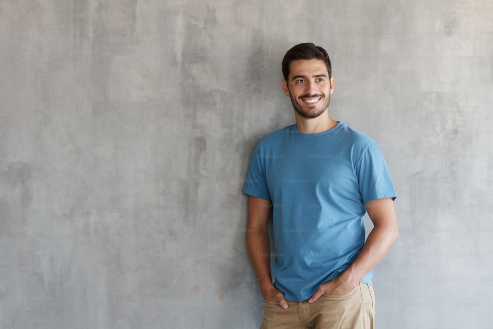Indoor picture of handsome Caucasian guy pictured isolated on gray background posing against gray textured wall, smiling happily, copyspace on left for promoting products for electronic commerce