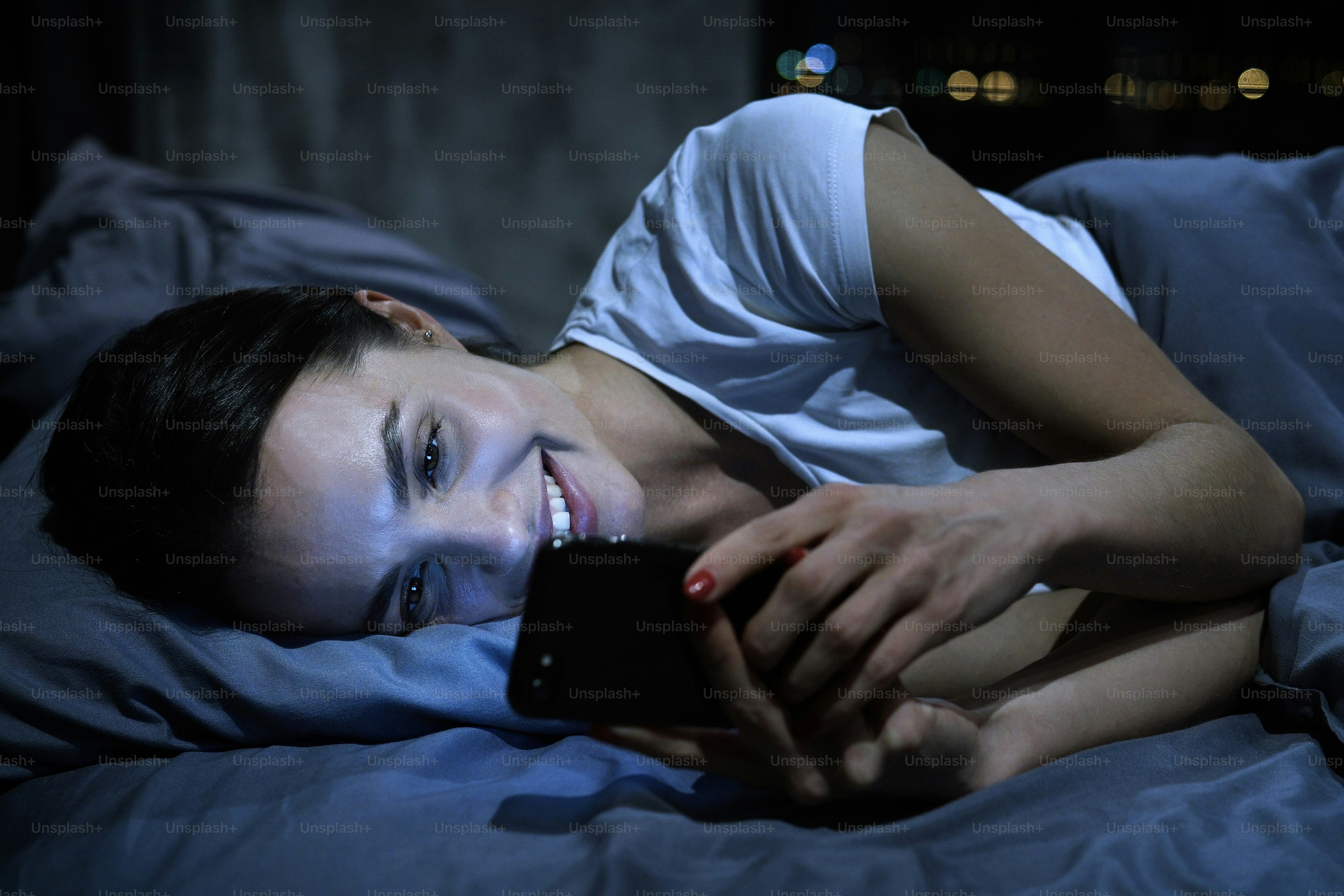 Young beautiful smiling female using smartphone in bed at night. Dating app concept.
