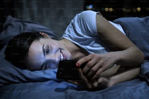 Young beautiful smiling female using smartphone in bed at night. Dating app concept.
