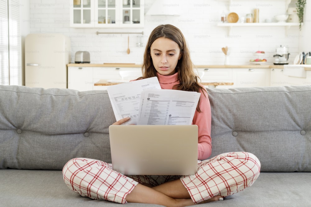 Young girl siiting on sofa in living room with laptop open and bills checking details of payment