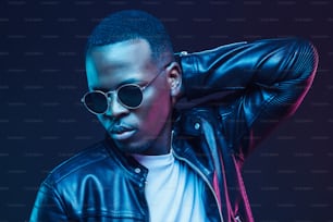 Neon light studio portrait of african american male model wearing trendy leather jacket and sunglasses