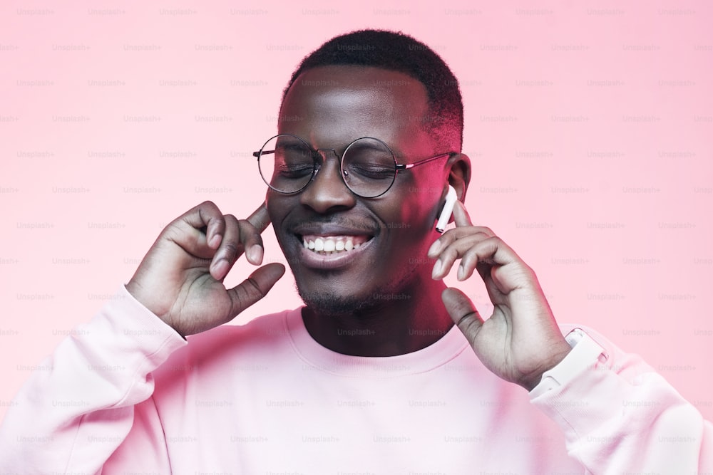 Foto de estudio de un joven africano escuchando música con auriculares inalámbricos aislados sobre fondo rosa