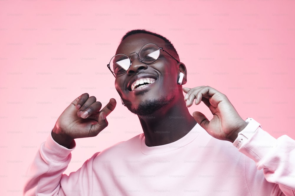 Young african man listening to music with earphones, dancing isolated on pink background