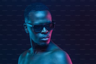 Retrato en primer plano de un hombre africano de pie con gafas de sol y con los hombros desnudos a la luz de neón
