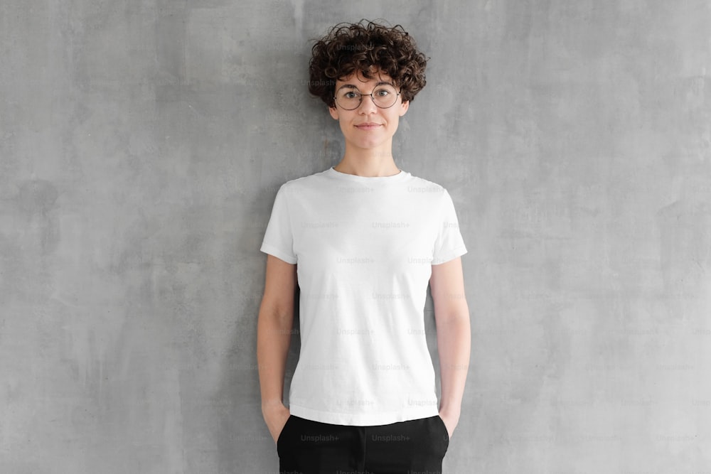텅 빈 흰색 면 티셔츠를 입고 회색 질감의 벽에 서서 포즈를 취하는 매력적인 젊은 여성