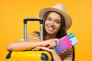 旅行のコンセプト。黄色い背景にパスポートと航空券とスーツケースを持つ若い幸せな女子学生旅行者