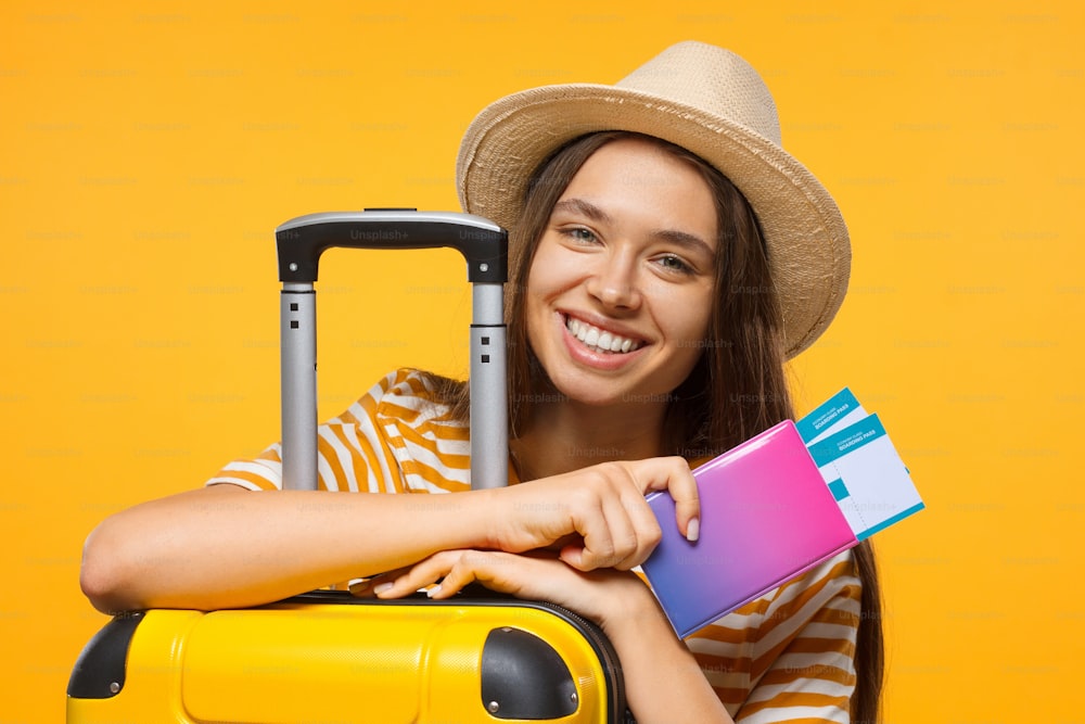 Travel concept. Young happy female student traveller holding passport with flight tickets and suitcase, isolated on yellow background