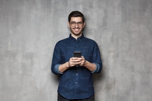 Moderner Geschäftsmann in Jeanshemd und trendiger Brille, der an einer grauen, strukturierten Wand steht und sein Telefon mit beiden Händen hält
