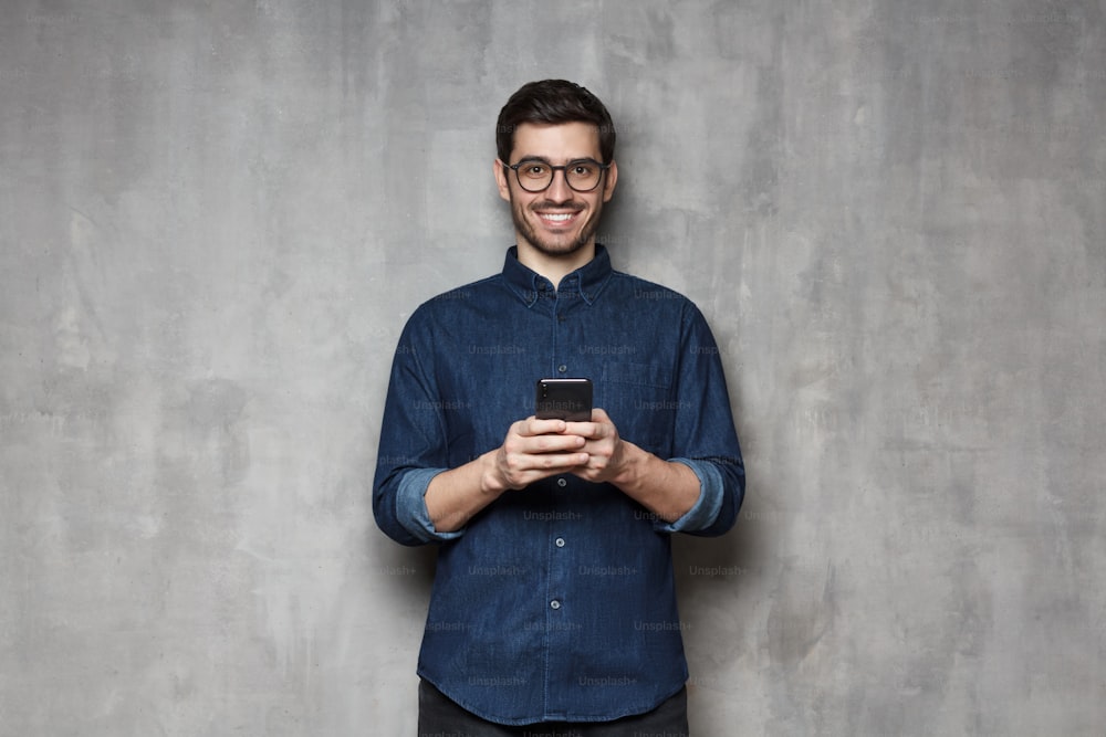 Homme d’affaires moderne souriant en chemise en jean et lunettes à la mode debout contre un mur texturé gris, tenant son téléphone à deux mains