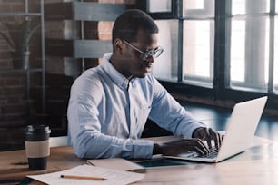 Portrait of young african business man sitting at his desk in loft office, typing message using laptop