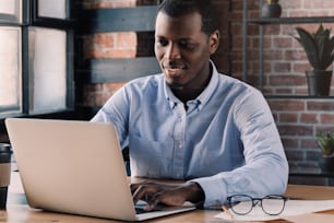 Close-up of positive african business man sitting in front of laptop in cafe against red brick wall