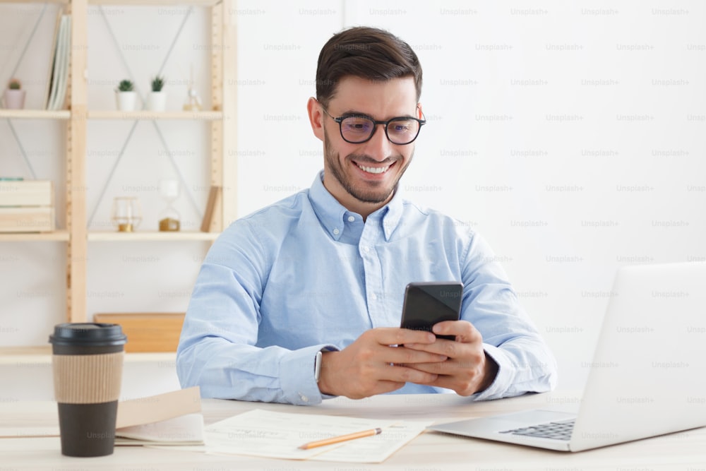 Young man wearing eyeglasses and shirt in office spending free time scrolling funny posts on his smartphone, smiling and laughing happily while looking at screen