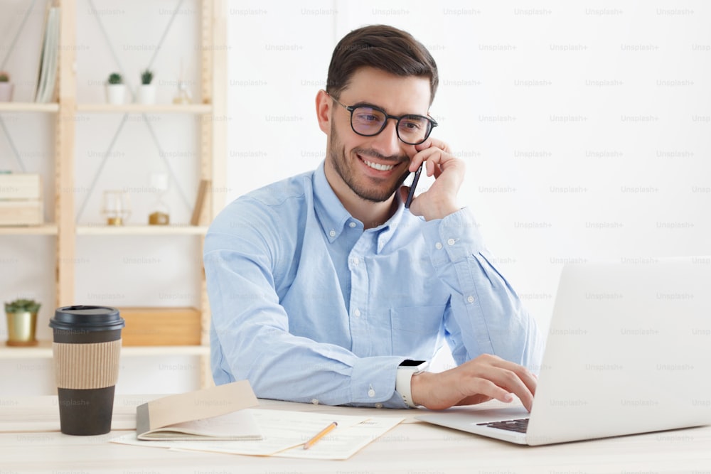 Young business man wearing glasses, working in office, looking attentively at screen, typing, answering phone call and consulting partner or client, smiling with confidence