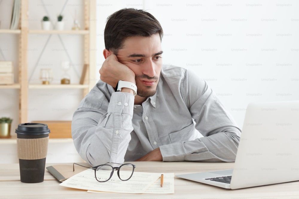 Homem de negócios em seu escritório trabalhando em laptop, descansando de cabeça na mão porque está cansado, fazendo horas extras de trabalho, estressado e entediado