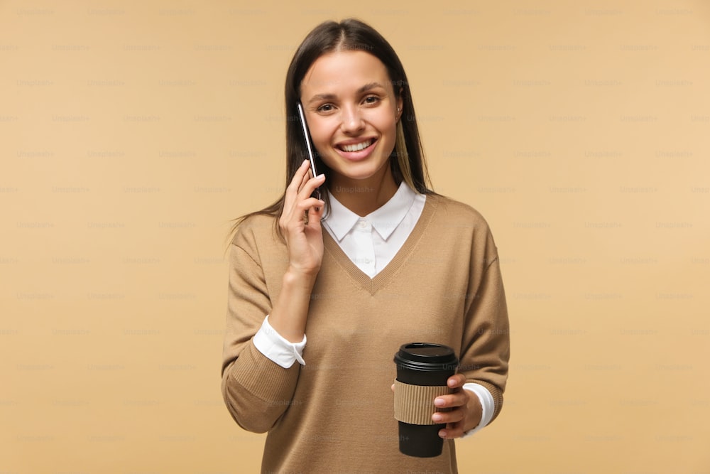 Retrato de estudio de una joven sonriente hablando por teléfono y sosteniendo café para llevar