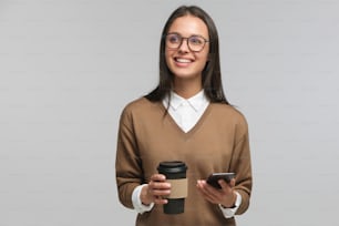 Young good-looking european woman isolated on gray background, holding phone and coffee to go