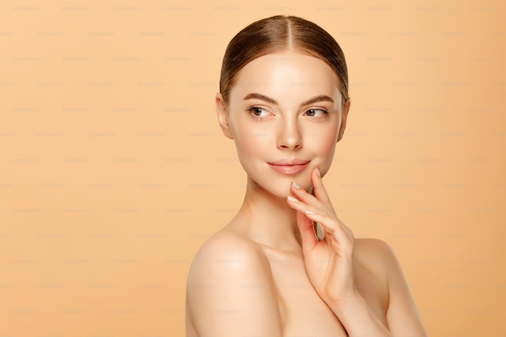 Young beautiful woman with brown eyes and glowing skin touching her face and looking away, isolated on beige background