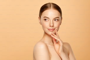 Young beautiful woman with brown eyes and glowing skin touching her face and looking away, isolated on beige background
