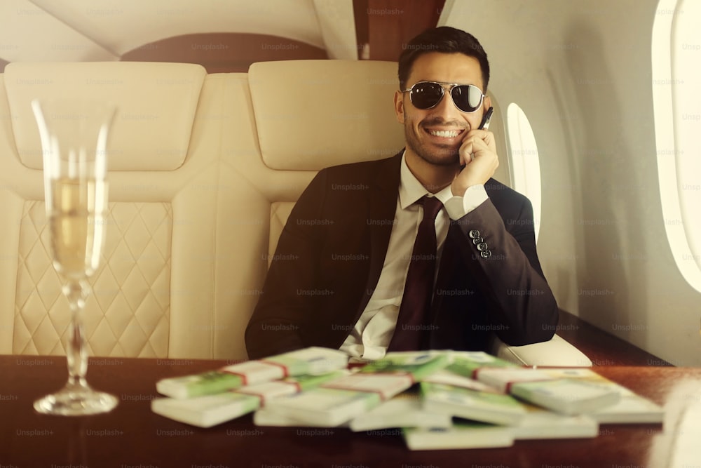 Giovane ricco felice a bordo del suo jet privato seduto a tavola con champagne ed euro in contanti, parlando al telefono