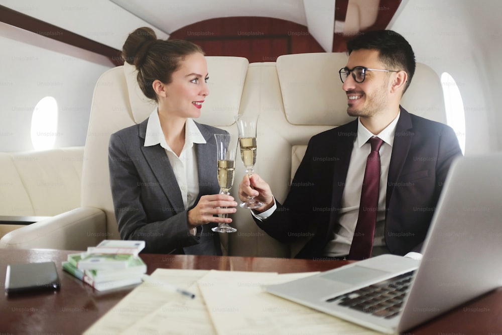 Business-Team aus jungen Männern und Frauen, die im Privatjet reisen, mit offenem Laptop und Geldscheinpackungen am Tisch sitzen und Champagner trinken, um Erfolge zu feiern