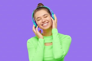 Menina sorridente feliz jovem vestindo neon verde manga comprida top, ouvindo música através de fones de ouvido sem fio com olhos fechados, isolado no fundo roxo