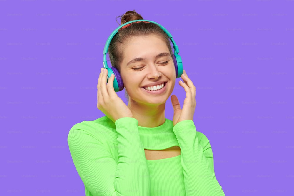 Happy smiling girl in neon green long sleeve top, listening to favorite music in wireless headphones, enjoying her free time, isolated on purple background