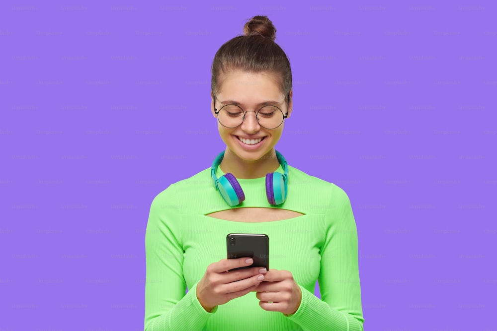 Young girl in glasses and neon top, with headphones around neck, holding phoen in hands, reading and texting messages with happy smile, isolated on purple background