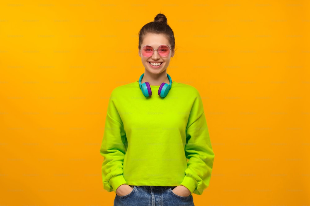 Junges Teenager-Mädchen trägt neongrünes Sweatshirt, blaue Jeans, rosa Brille und Kopfhörer um den Hals, steht mit den Händen in den Taschen, isoliert auf gelbem Hintergrund
