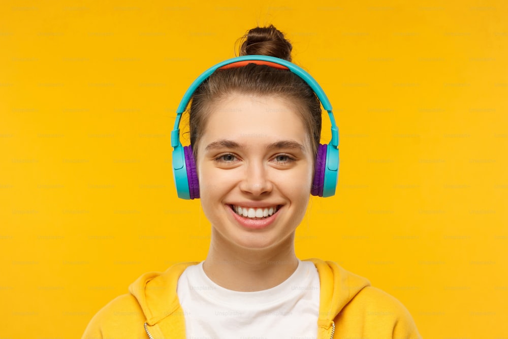 Jovem adolescente sorridente vestindo camiseta, capuz e fones de ouvido sem fio azuis, ouvindo música favorita e relaxando, isolado no fundo amarelo