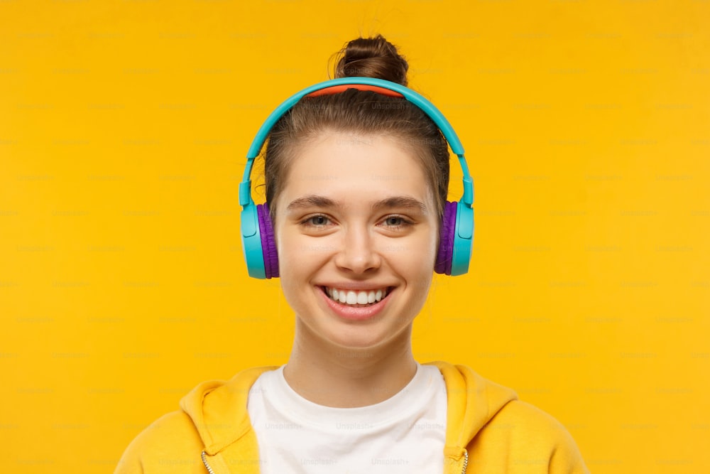 Young smiling teenage girl wearing t-shirt, hoodie and blue wireless headphones, listening to favorite music and relaxing, isolated on yellow background