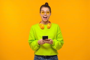 Retrato da mulher jovem vestindo óculos coloridos, moletom neon verde e fones de ouvido ao redor do pescoço, animado pelo conteúdo na tela do telefone, isolado no fundo amarelo