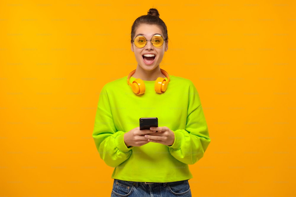Portrait of young woman wearing colored eyeglasses, green neon sweatshirt and headphones around neck, excited by content on phone screen, isolated on yellow background