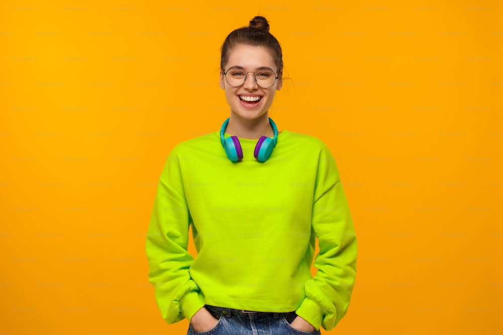 Young happy laughing girl in green neon sweatshirt and jeans, wearing blue wireless headphones around neck, keeoing hands in pockets, isolated on yellow background