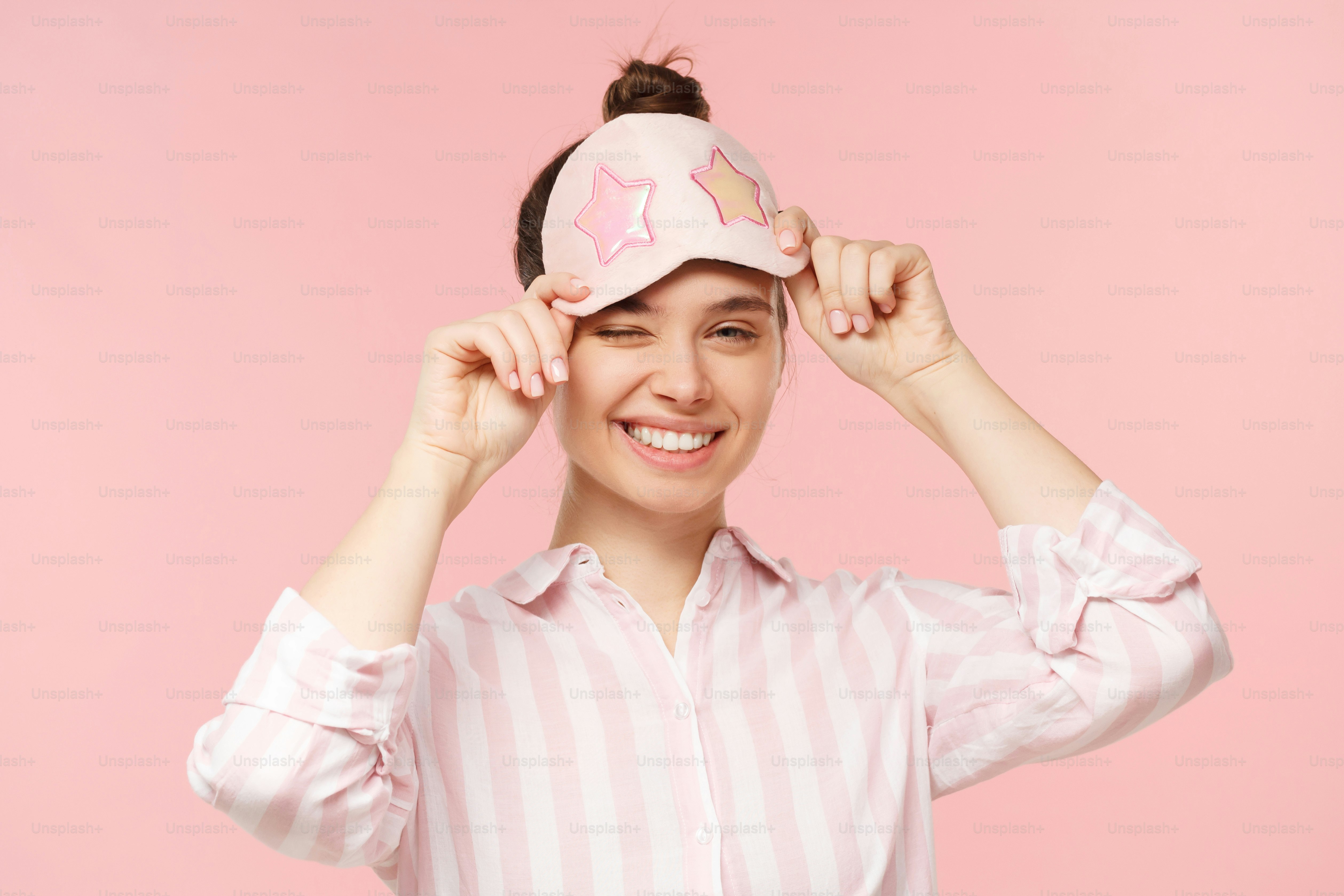 Joven mujer sonriente feliz con camisa a rayas y máscara para los ojos para dormir, manteniéndola en la frente y guiñando un ojo con un ojo, aislada sobre fondo rosa